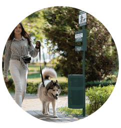 Pet Waste Station Installation. A woman walking her dog past a well-maintained pet waste station in a landscaped park, promoting responsible pet waste disposal.